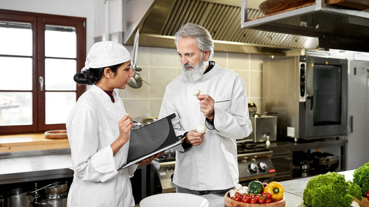 2 Chef sharing recipes. One is holding a cookbook and the other one is Teaching her how to cook.